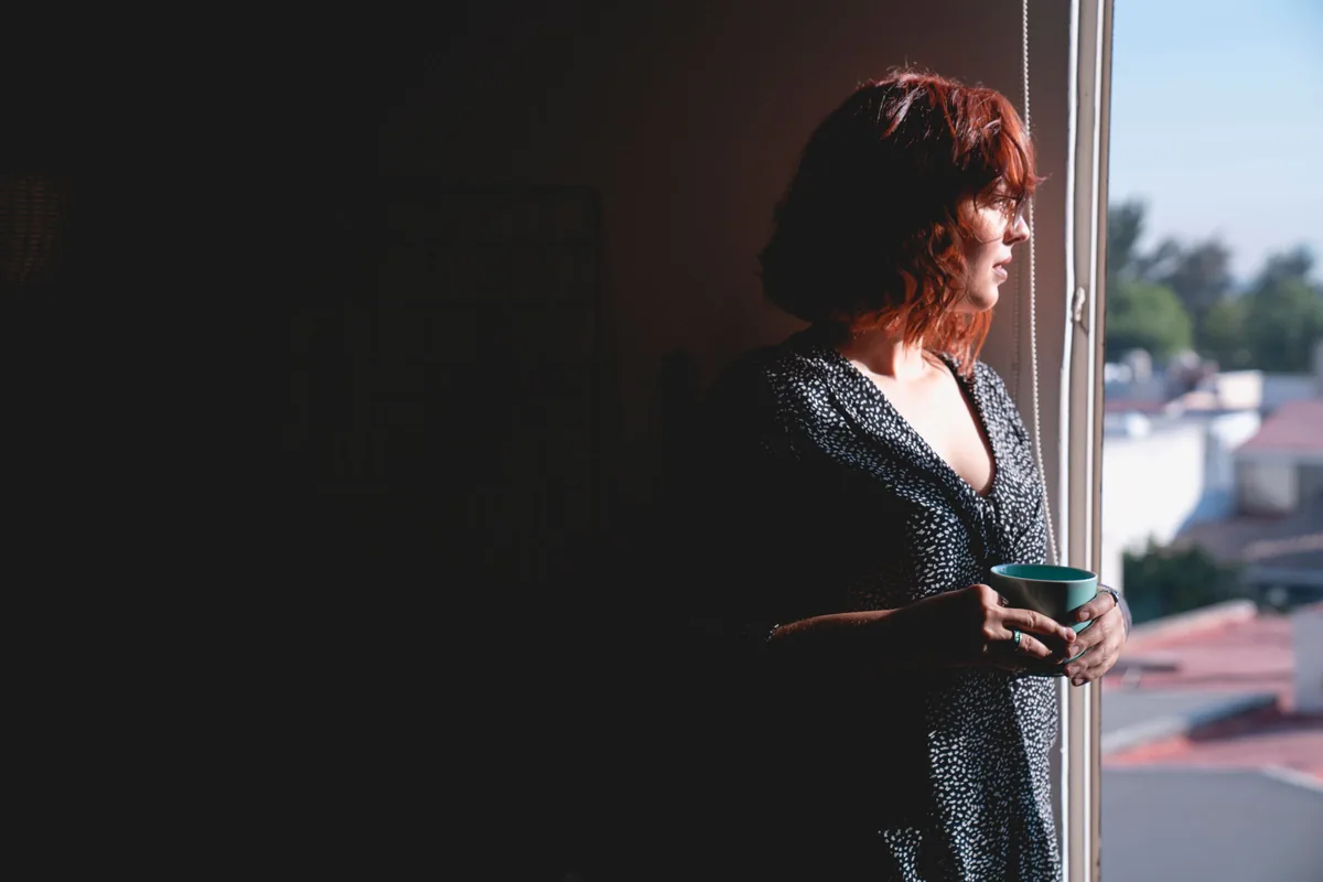 A woman holding a mug, looking out of a window, thinking something over